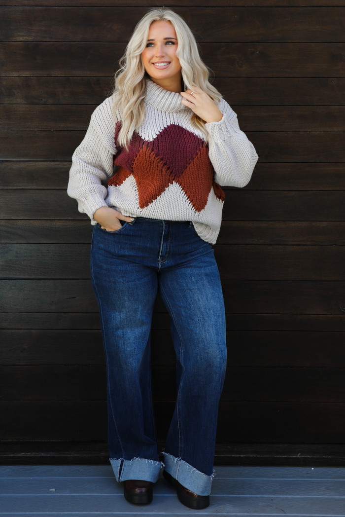 Woman model wearing a chunky knit turtle neck sweater with dark denim jeans outdoors in front of a dark wood door. 