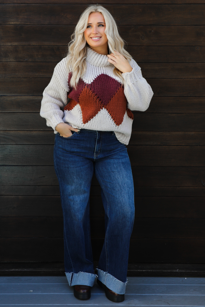 Woman model wearing a chunky knit turtle neck sweater with dark denim jeans outdoors in front of a dark wood door. 