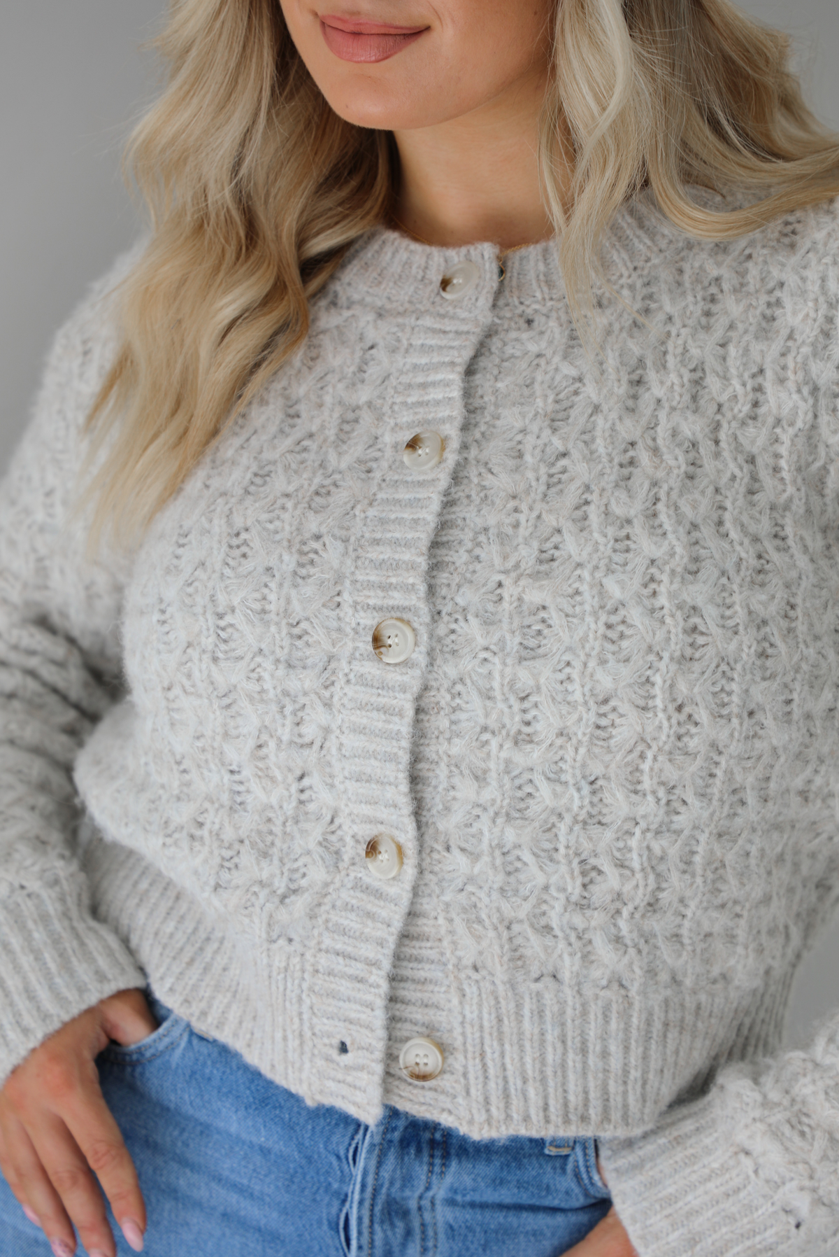 Woman wearing a light grey cardigan buttoned up with high waisted flared hem medium wash denim. The photo shows a closeup of the texture of the cardigan, which resembles a loose textured cable knit. The photo is taken indoors against a white wall.