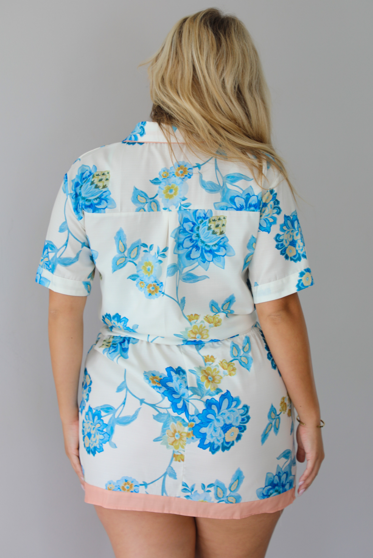 A woman shown from behind modeling a short-sleeve white button down top and skirt set with a blue floral print and a peach trim detail at the top, standing indoors against a white wall.