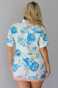 A woman shown from behind modeling a short-sleeve white button down top and skirt set with a blue floral print and a peach trim detail at the top, standing indoors against a white wall.