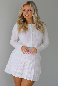 Woman wearing a two piece set that featuring a cropped cardigan and a high waisted skirt. The set is white with a pink horizontal striped pattern throughout, with scalloped hem details. The photo is taken indoors against a white wall.