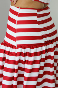 Woman wearing a red and white striped two piece set featuring a cropped top and maxi length, high waisted skirt. The photo is taken indoors against a white wall, and shows a closeup of the side of the skirt, which has  a zipper closure. 