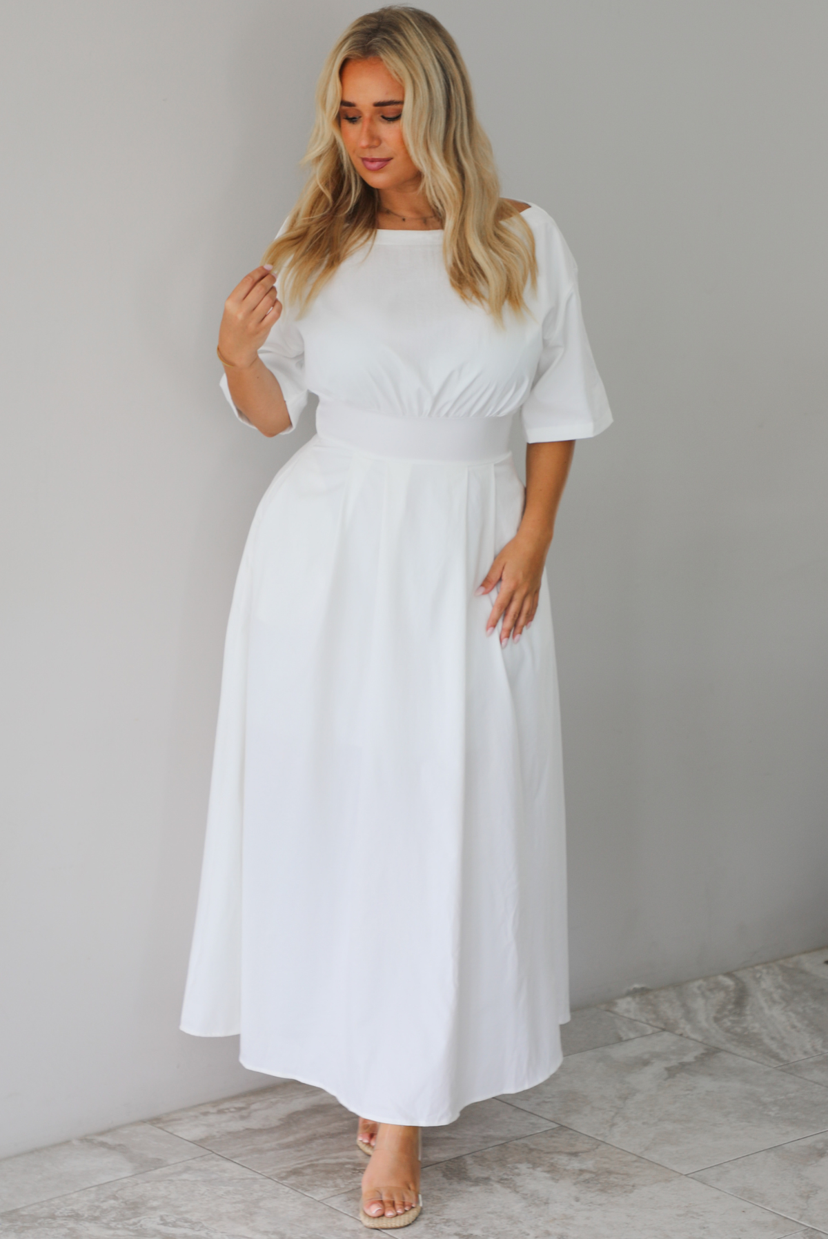 A woman modeling a white midi dress with short puff sleeves, a wide boat neckline, and side pockets, standing indoors against a white wall with white marble flooring. 