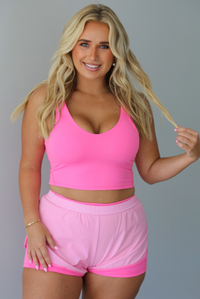 A woman modeling a bright pink two-piece activewear set consisting of a scoop-neck crop top and matching athletic shorts with a darker, hot pink trim, standing indoors against a white wall.
