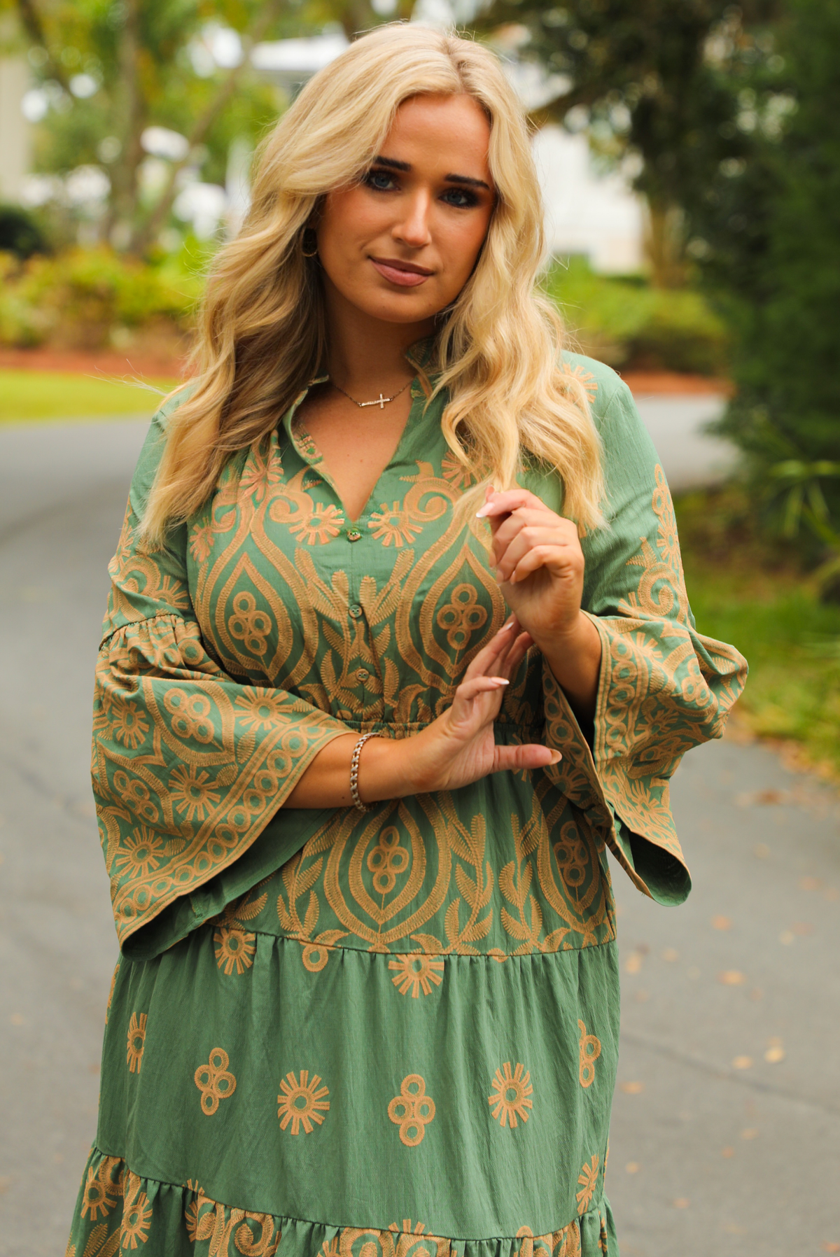 Woman wearing a teal dress that has tan a stitched pattern throughout, quarter length  sleeves, a collared, v-neckline, cinched with elastic waistline, and a maxi-length skirt. The photo shows a close up of the bodice of the dress which has non-functional button closures, and is taken outdoors standing on a paved road with trees and other greenery in the background.
