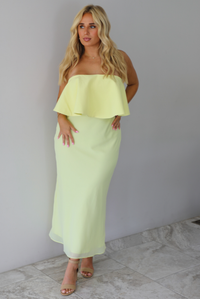A woman modeling a pale yellow strapless midi dress with a ruffled detail at the top that covers the bodice and a fitted skirt, standing indoors against a white wall with white marble flooring. 