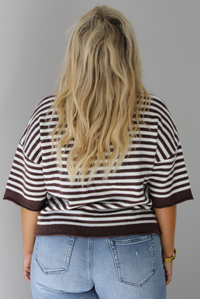 A woman shown from behind wearing a brown and white striped knit button top cardigan top, paired with light wash denim distressed denim, standing indoors against a white wall.