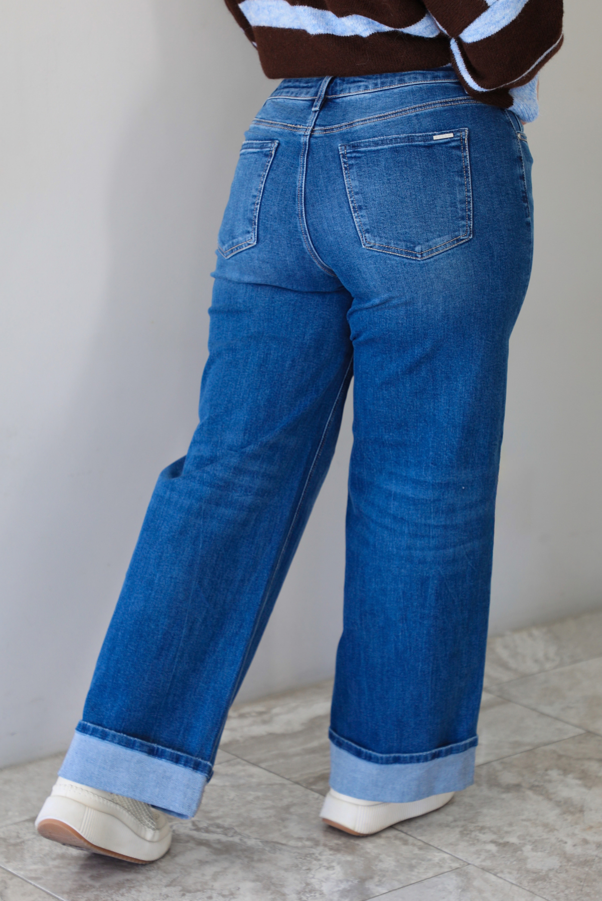 A closeup of a woman shown from behind wearing dark denim jeans with a cuffed hem, standing indoors against a white wall with white marble flooring. 