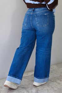 A closeup of a woman shown from behind wearing dark denim jeans with a cuffed hem, standing indoors against a white wall with white marble flooring. 