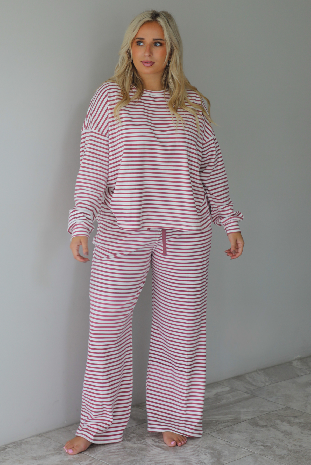 A woman wearing a matching two piece set featuring a long sleeved top and high waisted wide leg pants with a berry and white striped pattern throughout, standing indoors against a white wall with white marble flooring. 
