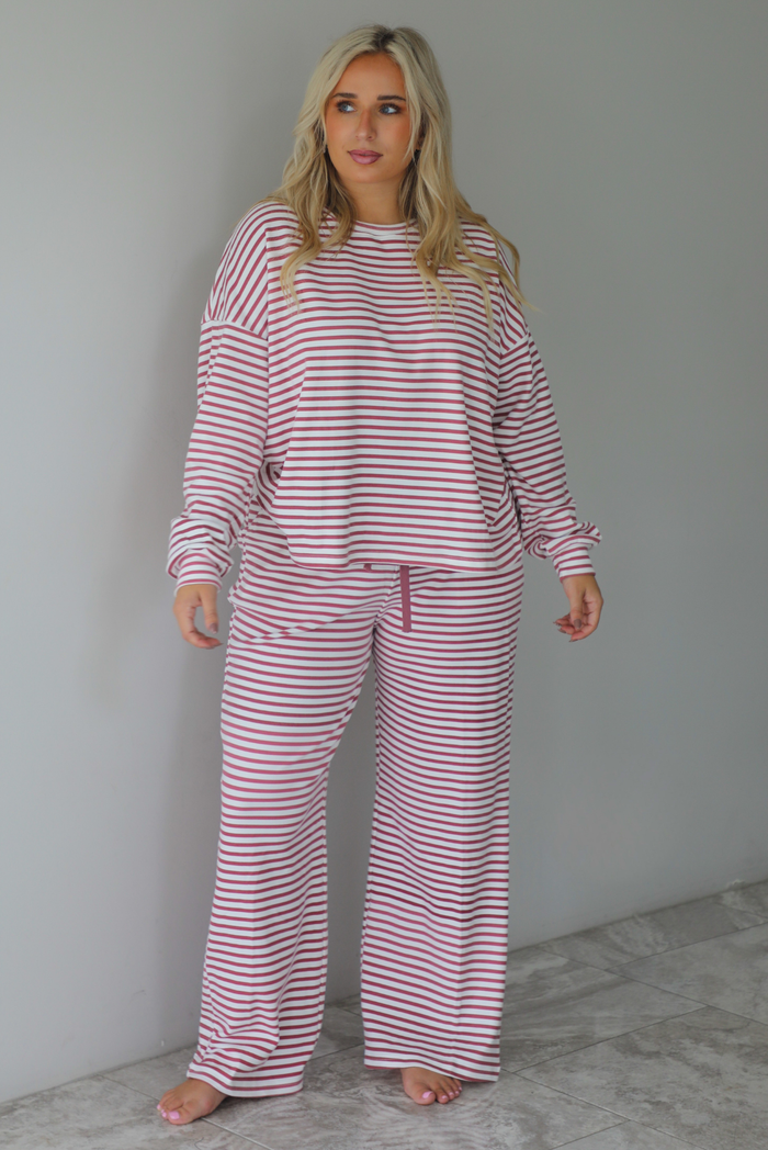 A woman wearing a matching two piece set featuring a long sleeved top and high waisted wide leg pants with a berry and white striped pattern throughout, standing indoors against a white wall with white marble flooring. 
