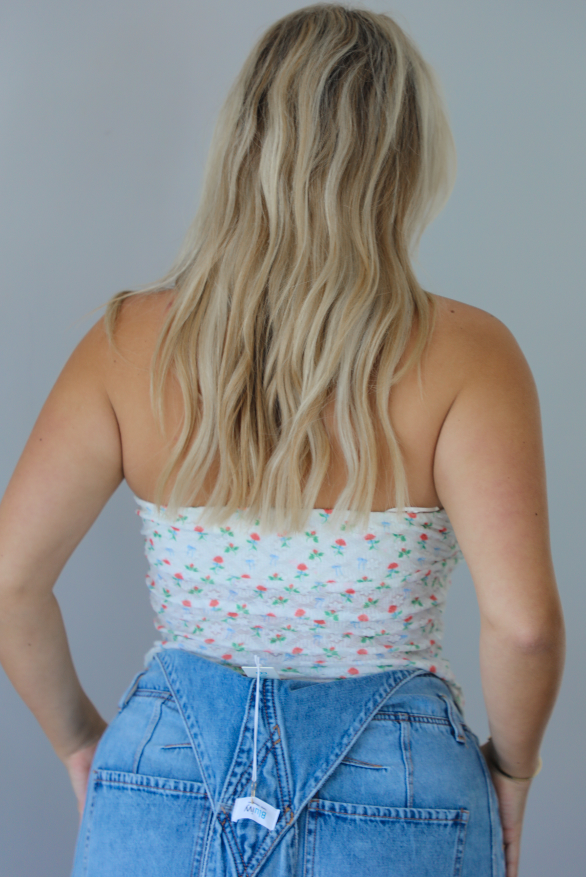 Woman wearing a floral strapless top and denim overalls indoors against a white wall. The photo shows the back of the top.