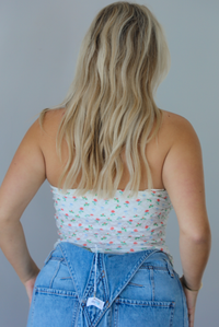 Woman wearing a floral strapless top and denim overalls indoors against a white wall. The photo shows the back of the top.