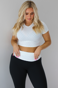 Woman wearing a two piece set featuring a white cropped top with a black polka dot print pattern and black pants that are high waisted with a white and black polka dot print folded over detail. The photo is taken indoors against a white wall. The photo shows a closeup of the top. 