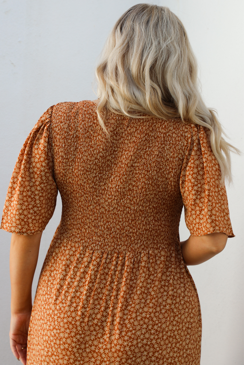 Up close of the back of a woman model outdoors wearing a rust dress with a cream floral print pattern throughout. It has flowy short sleeves, a smocked bodice, and a flowy maxi length skirt with functional pockets on either side. 