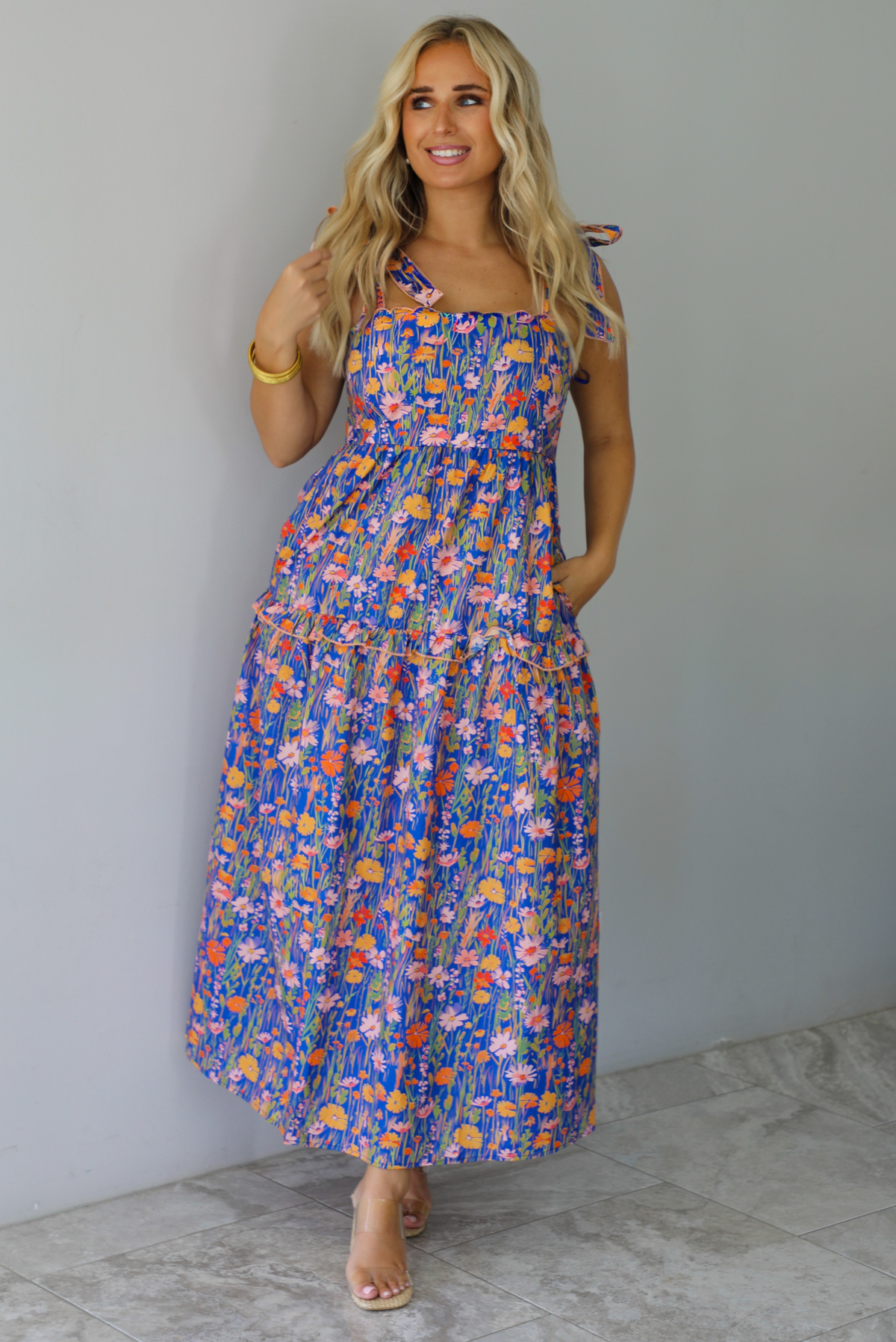 A woman wearing a sleeveless tiered navy blue maxi dress with a colorful floral print with a fitted bodice and flowy skirt with functional pockets on either side, standing indoors against a white wall with white marble flooring. 