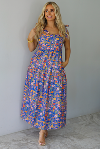 A woman wearing a sleeveless tiered navy blue maxi dress with a colorful floral print with a fitted bodice and flowy skirt with functional pockets on either side, standing indoors against a white wall with white marble flooring. 
