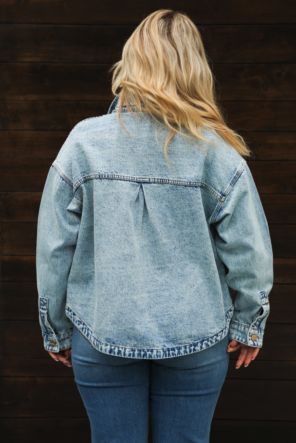 Woman model wearing an oversized, acid washed, light denim jacket, paired with a sheer mesh leopard print top, and medium wash denim jeans. The photo shows the back of the jacket.
