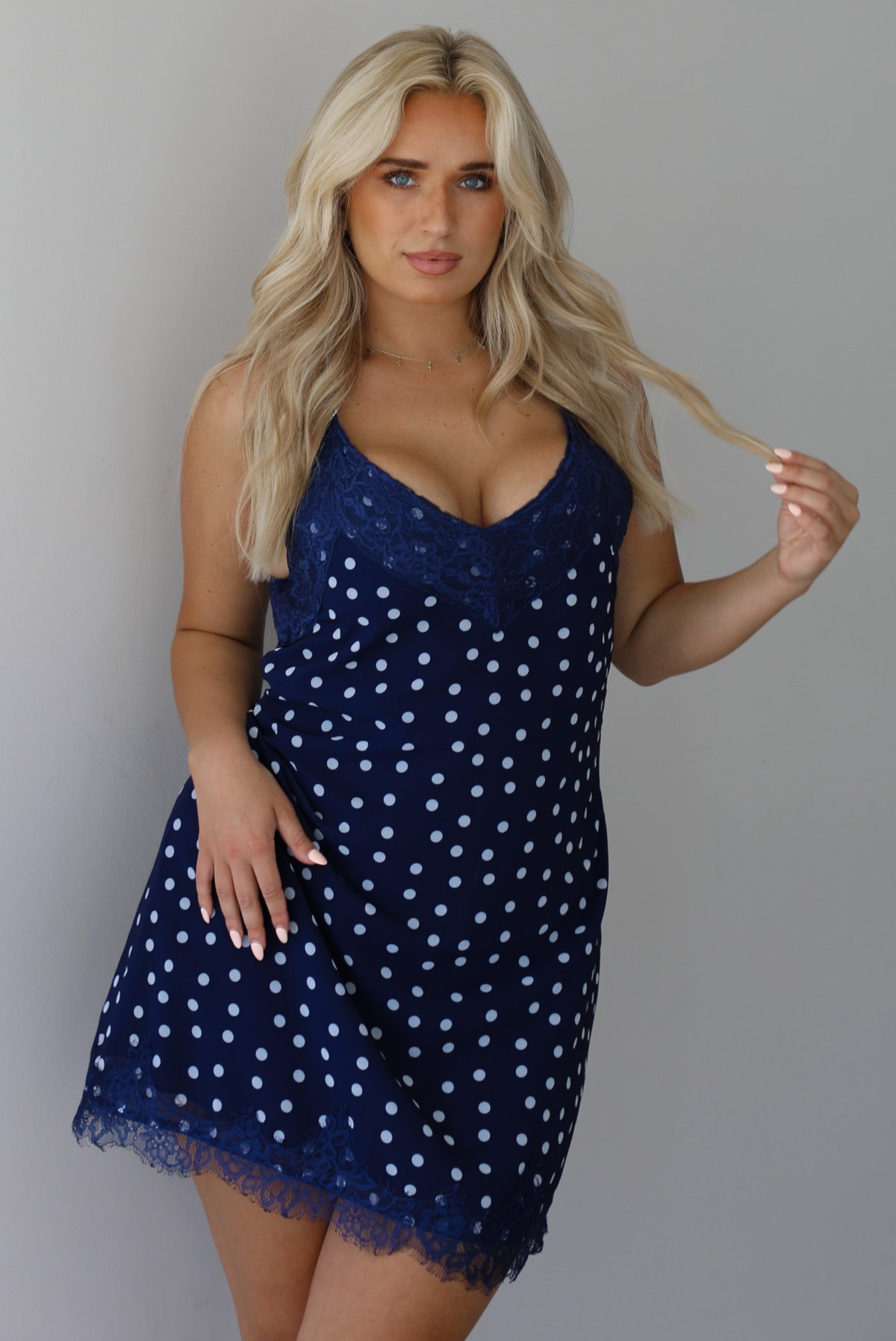 Woman wearing a navy dress with a white polka dot print pattern throughout with blue lace detailing at the neckline and hem indoors against a white wall.
