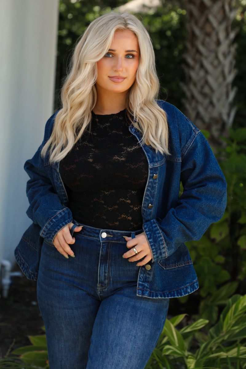 Close up of a woman model wearing a dark denim button up jacket with a black lace top,  and dark denim jeans outdoors. 