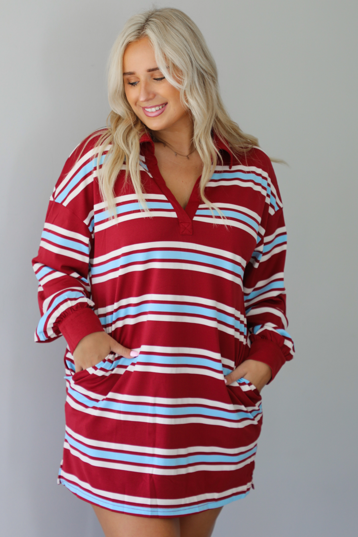A woman wearing a red burgundy mini dress with a white and blue striped pattern throughout, collared neckline, long sleeves, and functional pockets on ether side, standing indoors against a white wall.