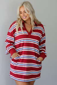 A woman wearing a red burgundy mini dress with a white and blue striped pattern throughout, collared neckline, long sleeves, and functional pockets on ether side, standing indoors against a white wall.