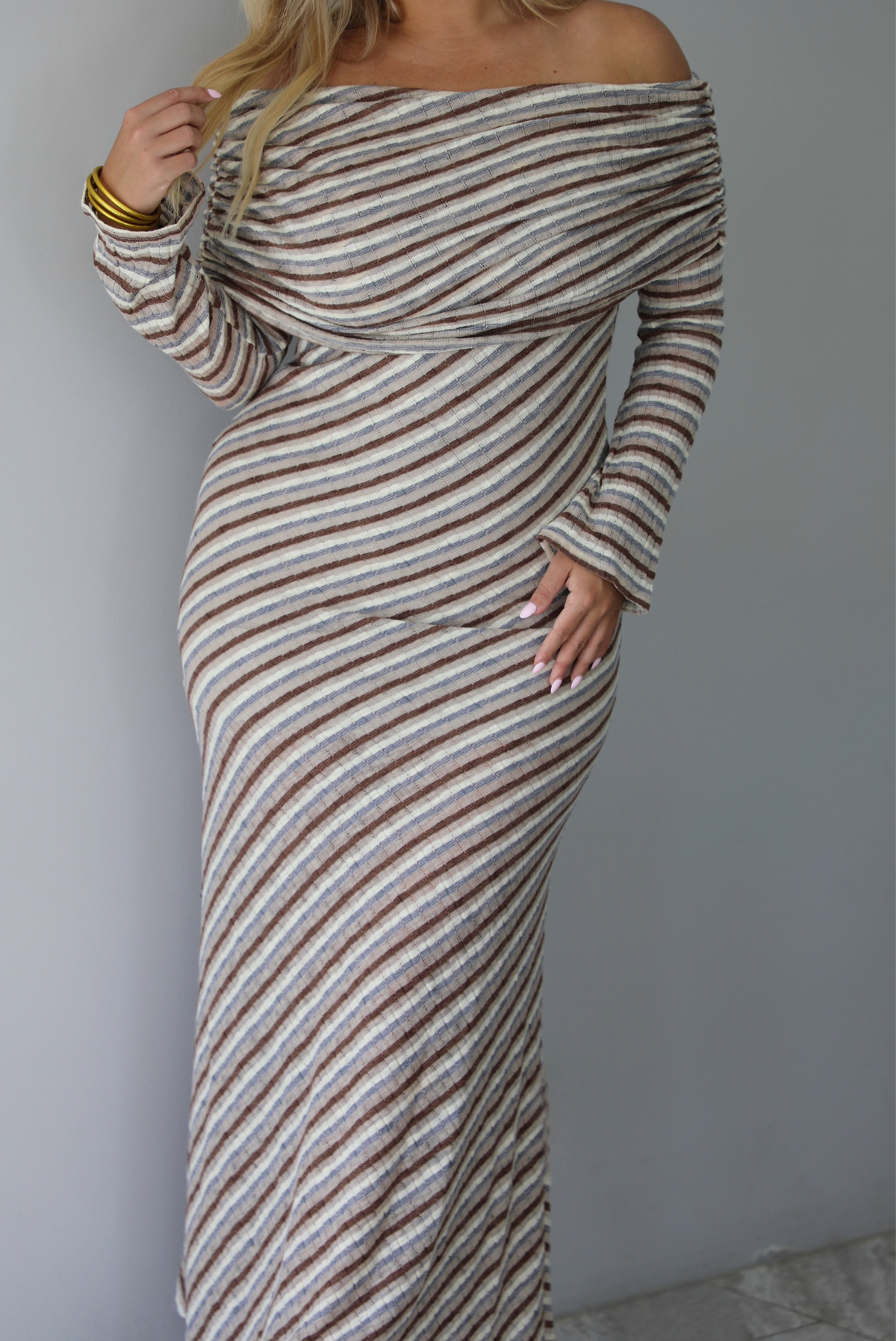 Woman wearing a grey, brown, muted blue, and off white striped fitted maxi dress that has a folded down design at the shoulders and bodice and long sleeves, standing indoors against a white wall. The photo shows a closeup of the dress without showing the models face or the hem of the dress. 
