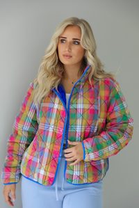 Woman wearing a multi-colored plaid, quilted style jacket paired with a white top underneath and high waisted blue lounge pants. The photo shows a closeup of the jacket, with the functional button closures in the front, functional pockets, and quilted texture. The photo is taken indoors against a white wall.