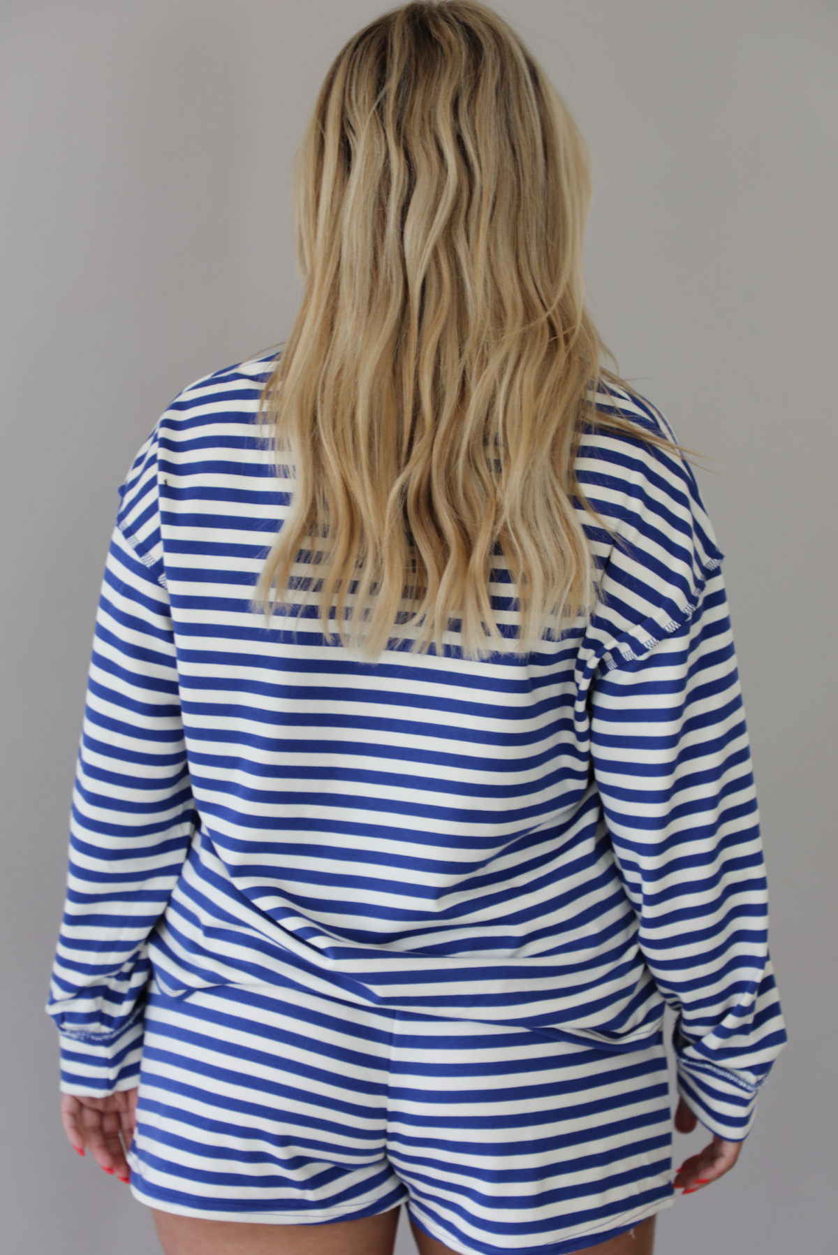 Woman wearing a navy and white striped two piece set featuring a long sleeved top and shorts indoors against a white wall. The photo shows the back of the set. 