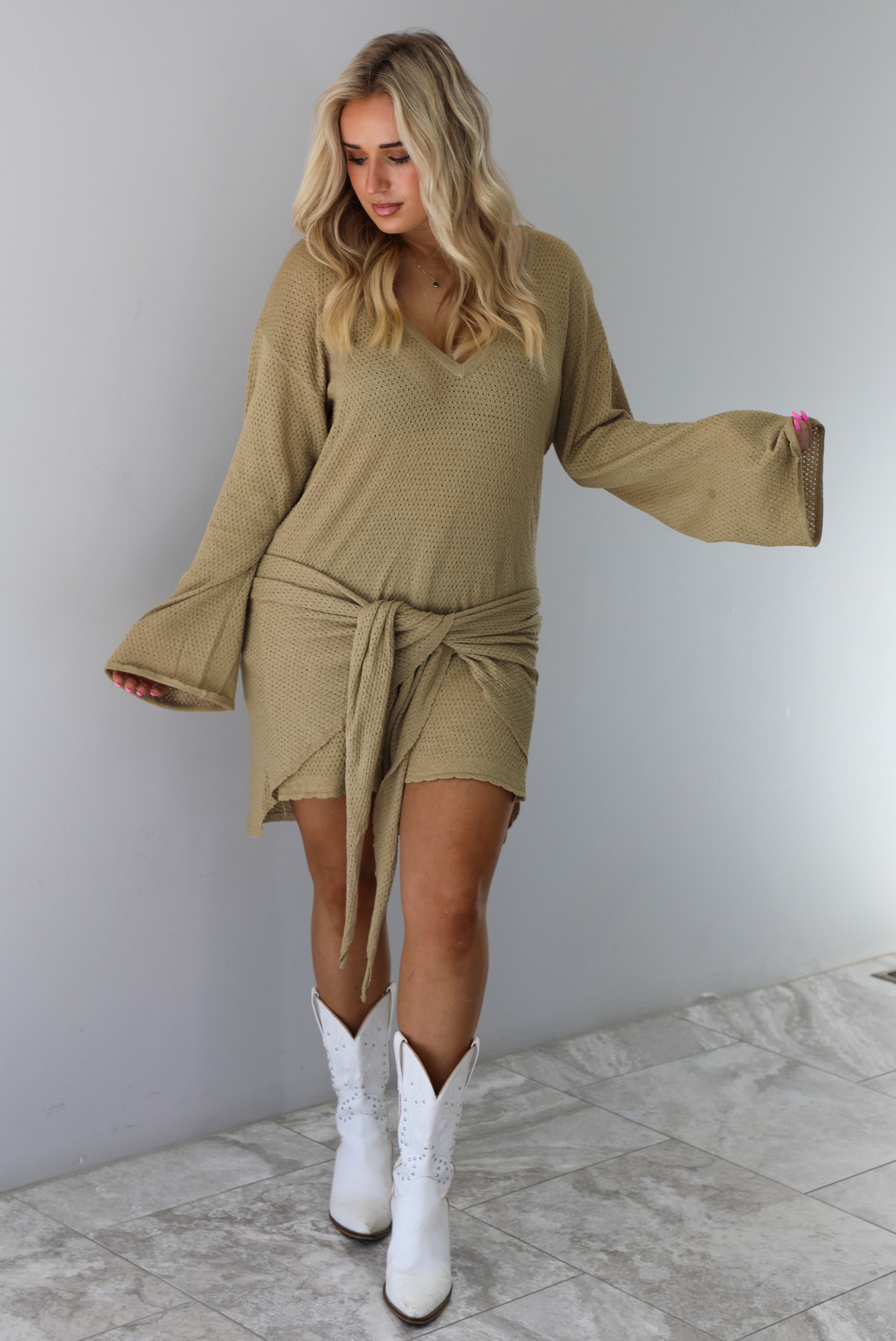Woman wearing a camel long-sleeve dress that has a tie detail in the front. The dress is paired with white cowboy boots. The photo is taken indoors against a white wall with white marble flooring. 
