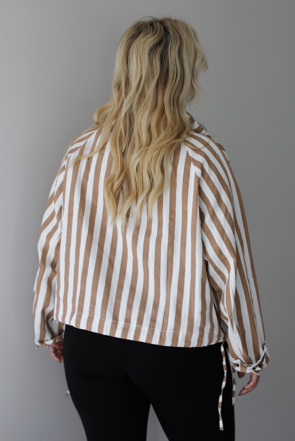 A woman shown from behind wearing a tan and white striped collared long sleeved top with functional button closures along the front, worn with black leggings, standing indoors against a white wall. 