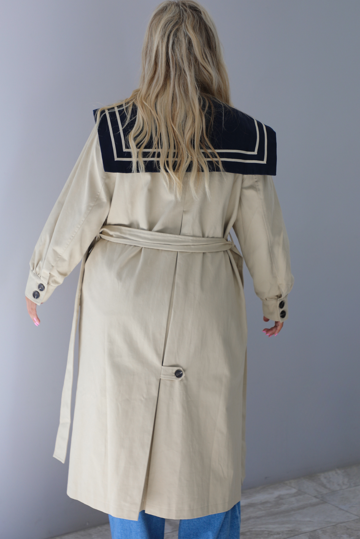 A woman shown from behind wearing a tan trench coat with a navy collared neckline, functional button closures in the front, functional pockets on either side, with a tie waistline, paired with light wash flared hem denim jeans, standing indoors against a white wall.