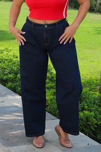 A woman wearing wide-leg dark denim jeans, a red crop top, and brown woven flat shoes, standing outdoors on a stone path with green grass and bushes in the background.