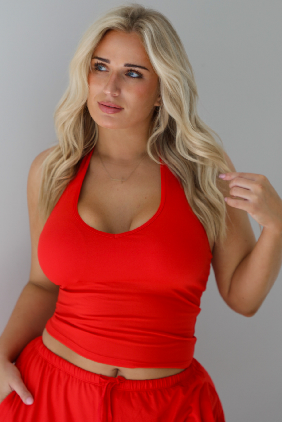 Woman wearing a red two piece set featuring a  tank top and slouchy pants. The photo shows a closeup of the tank top and the upper portion of the pants. The photo is taken indoors against a white wall.