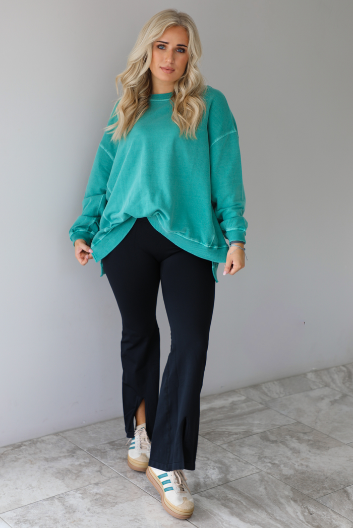 Woman wearing a teal oversized sweater with black leggings that have a slightly flared hem with split hem details in the front. The photo is taken in front of a white wall with white marble flooring. 