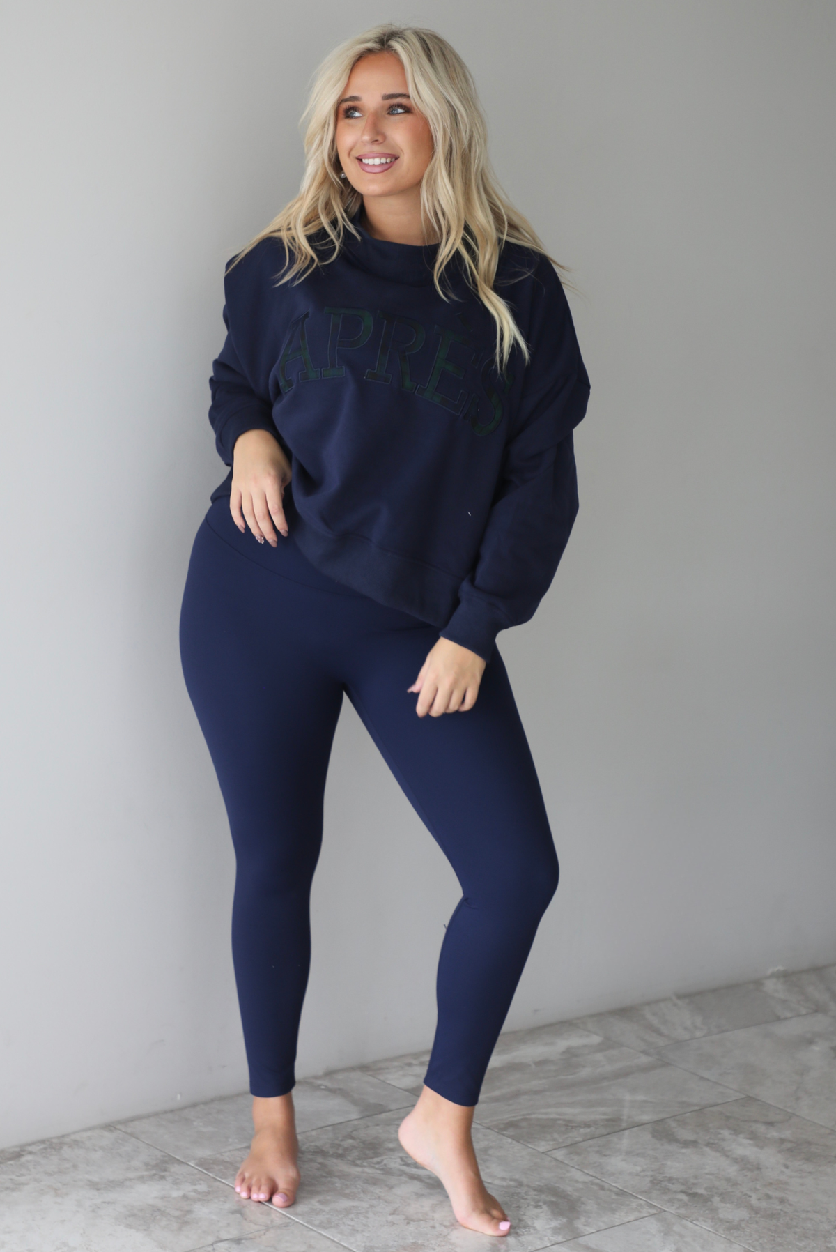 A woman wearing a navy sweater with Apreś written on the front in collegiate lettering in a plaid print, paired with a matching two piece navy set, standing indoors against a white wall with white marble flooring. 