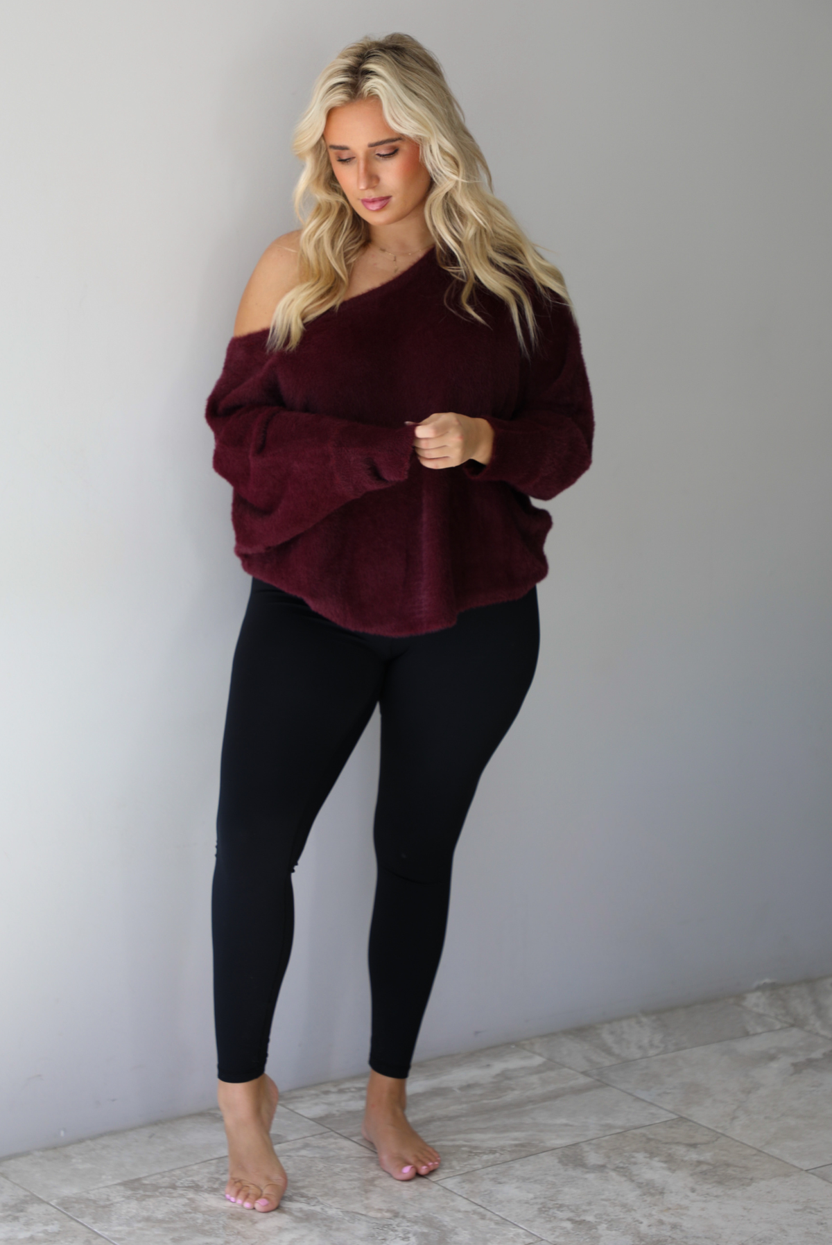 A woman wearing a maroon oversized long sleeved sweater, worn off the shoulder, paired with black leggings, indoors against a white wall with white marble flooring.