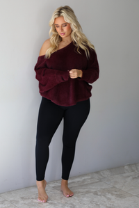 A woman wearing a maroon oversized long sleeved sweater, worn off the shoulder, paired with black leggings, indoors against a white wall with white marble flooring.