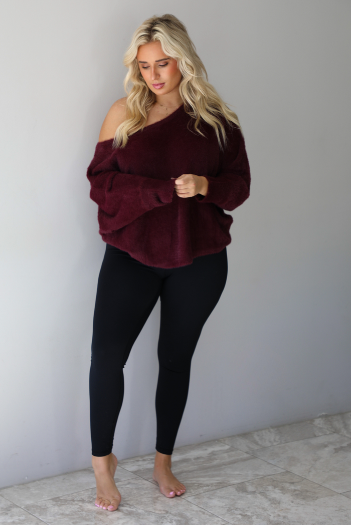 A woman wearing a maroon oversized long sleeved sweater, worn off the shoulder, paired with black leggings, indoors against a white wall with white marble flooring.
