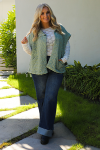 Woman model wearing a sage colored oversized vest paired with a white lace long sleeved top and dark denim outdoors.  The vest has functional pockets on either side, functional button closures in the front, and an oversized fit. 