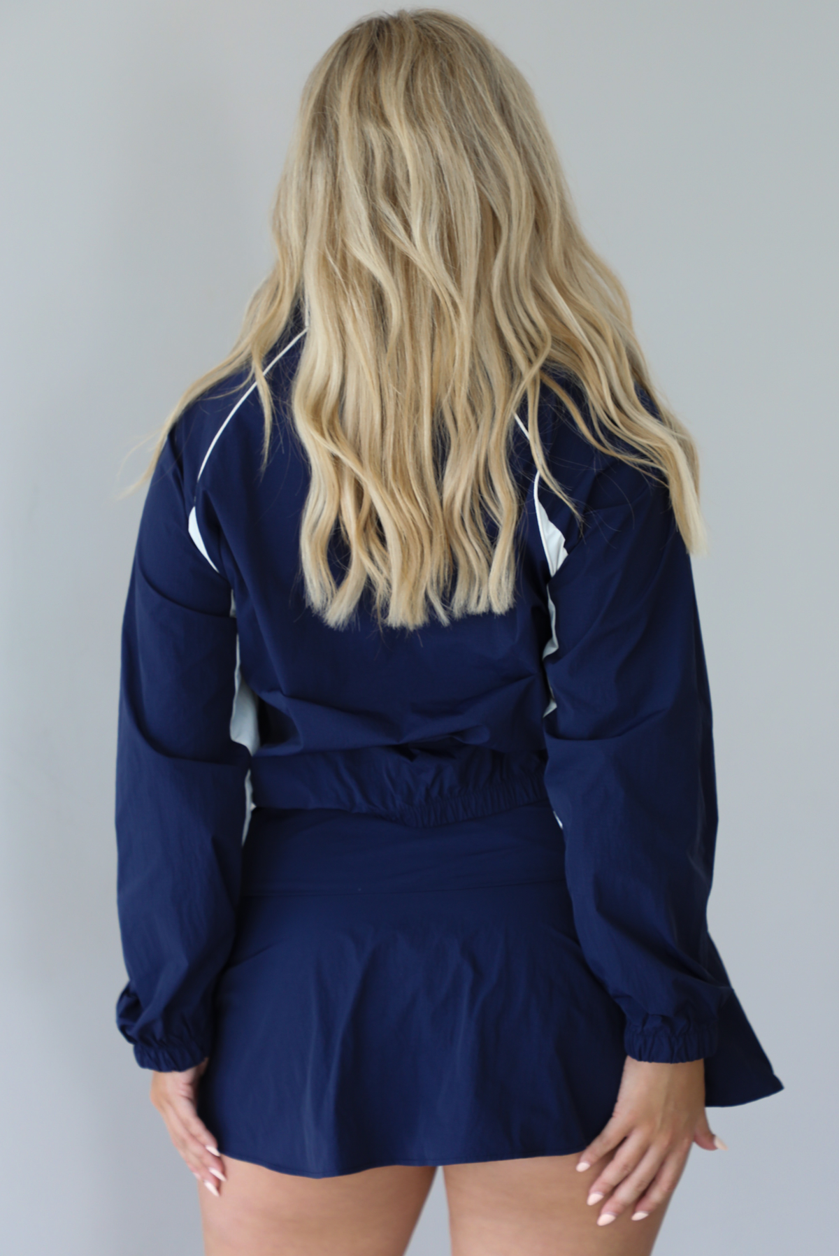 Woman wearing a navy tracksuit featuring a jacket and skort with a white tank indoors against a white wall. The photo shows the back of the set. 
