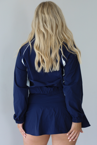 Woman wearing a navy tracksuit featuring a jacket and skort with a white tank indoors against a white wall. The photo shows the back of the set. 