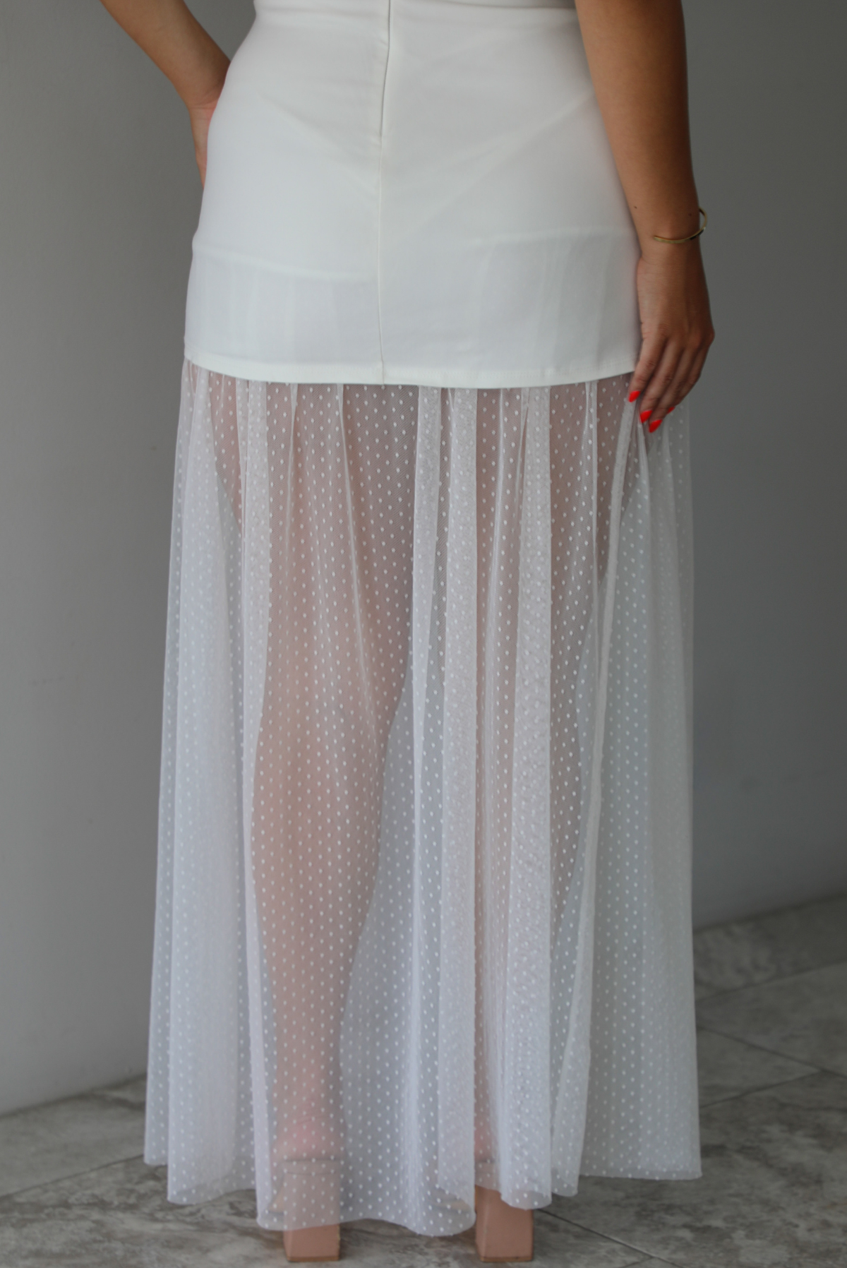 Woman wearing a white dress that is strapless, has a mini length, with mesh detailing giving it a maxi length indoors against a white wall with white marble flooring. The photo shows a close-up of the back of the dress, showing the sheer mesh hem of the dress and zipper closure in the back.