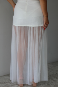 Woman wearing a white dress that is strapless, has a mini length, with mesh detailing giving it a maxi length indoors against a white wall with white marble flooring. The photo shows a close-up of the back of the dress, showing the sheer mesh hem of the dress and zipper closure in the back.