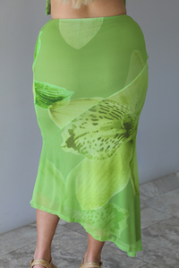 Woman wearing a green tropical print set featuring a backless tank and high waisted, asymmetrical hem midi skirt indoors against a white wall with white marble flooring. The photo shows a closeup of the back of the skirt. 