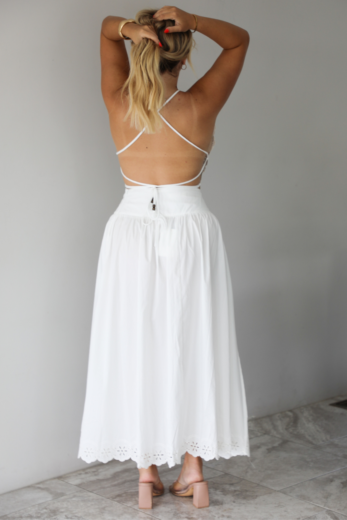 Woman wearing a two piece white set indoors against a white wall with white marble flooring. The photo shows the back of the set, which has an open back on the top. 