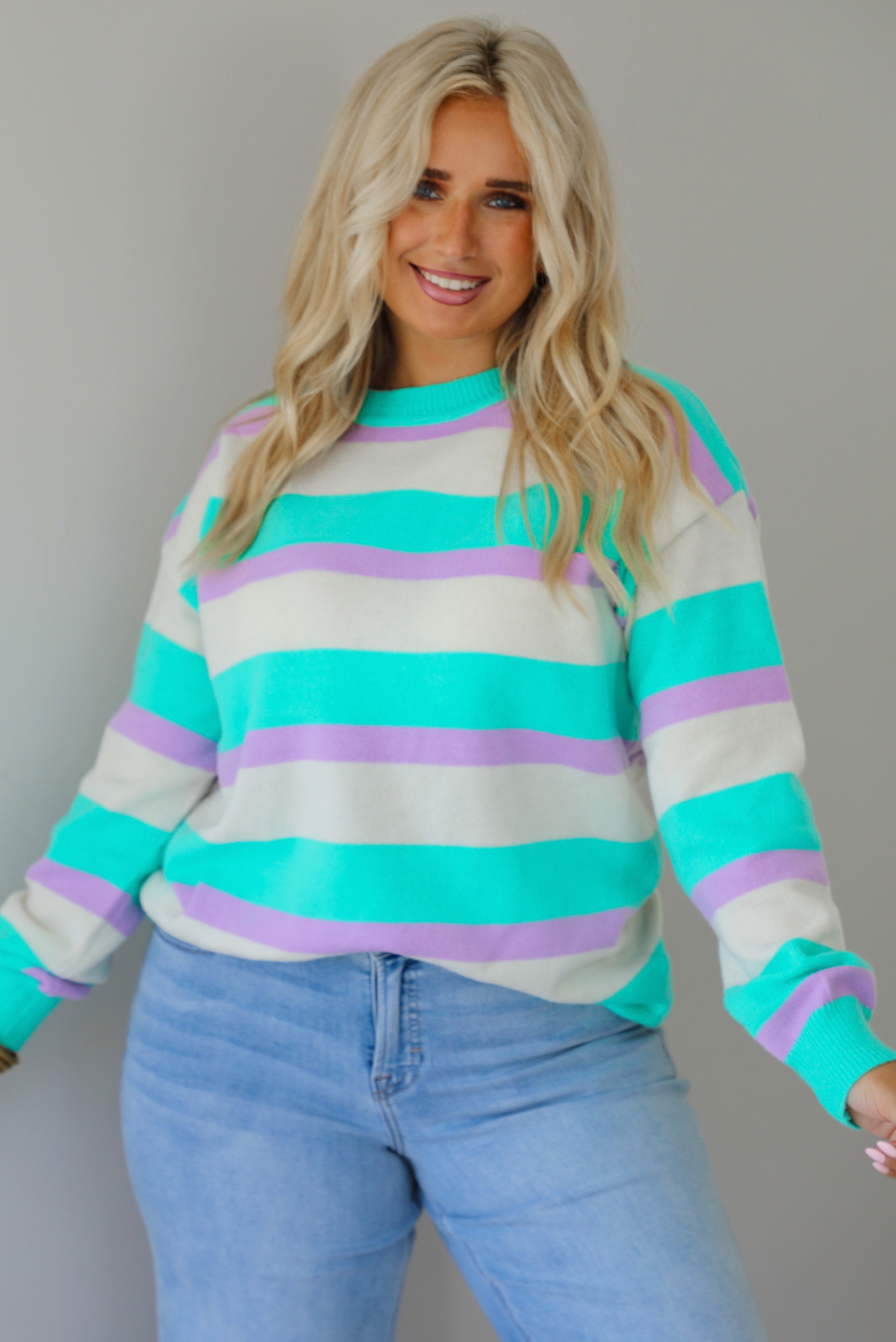 A woman wearing a turquoise, lavender, and cream striped knit sweater, paired with light wash denim jeans, standing indoors against a white wall.