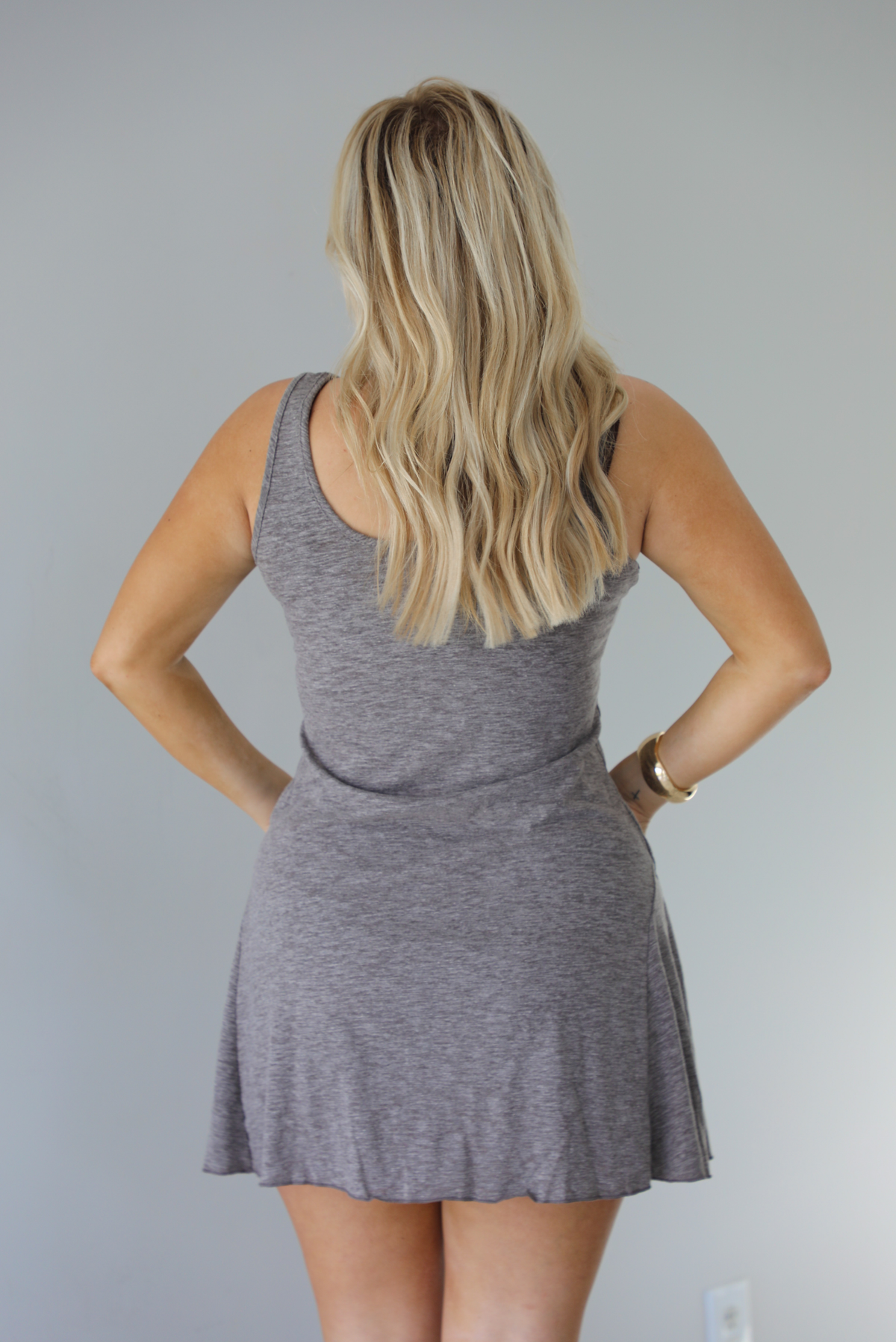 Woman wearing a charcoal dress that has thick, tank-style straps, exposed seams details, has a lightweight t-shirt textured material, and has a mini dress length. The photo is taken indoors against a white wall and shows the back of the dress. 