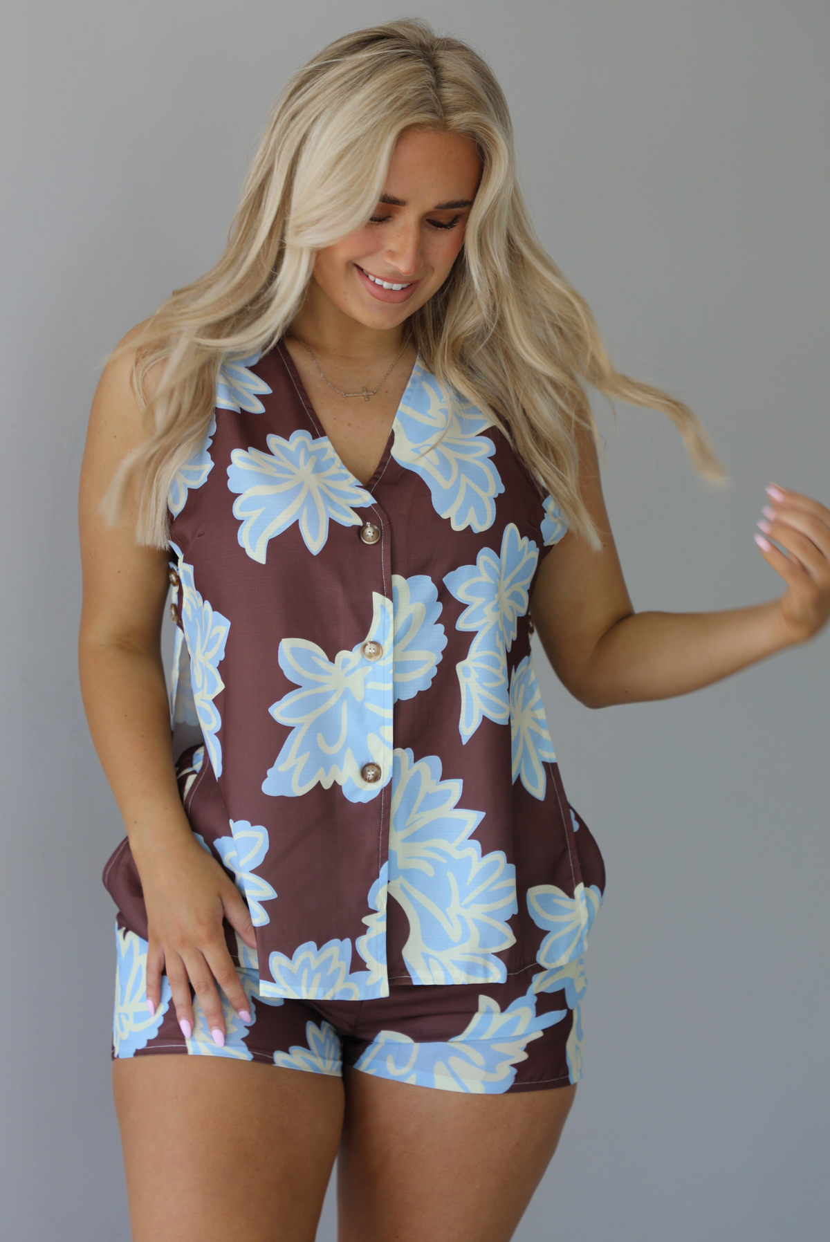 Woman wearing a brown, blue, and off white floral two piece set featuring a vest style top with functional button closures in the front and shorts. The photo is taken in front of a white wall.